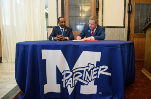 Chancellor signing paper with seated man, blue cloth says MPartner