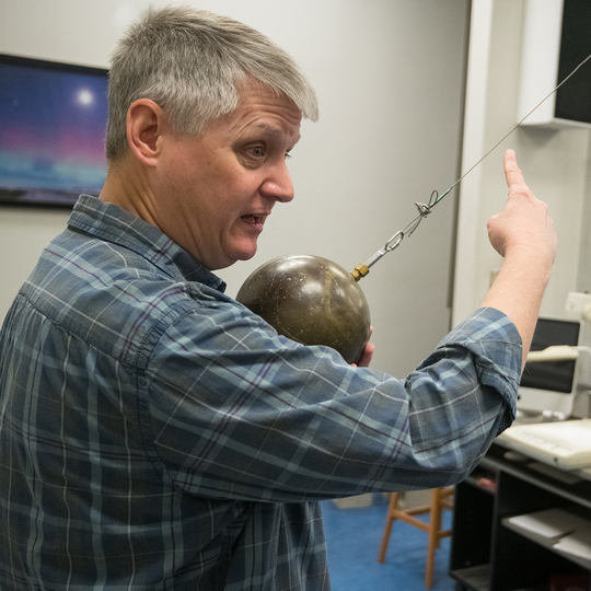 Breese Quinn in physics classroom holding a ball on a metal cord 